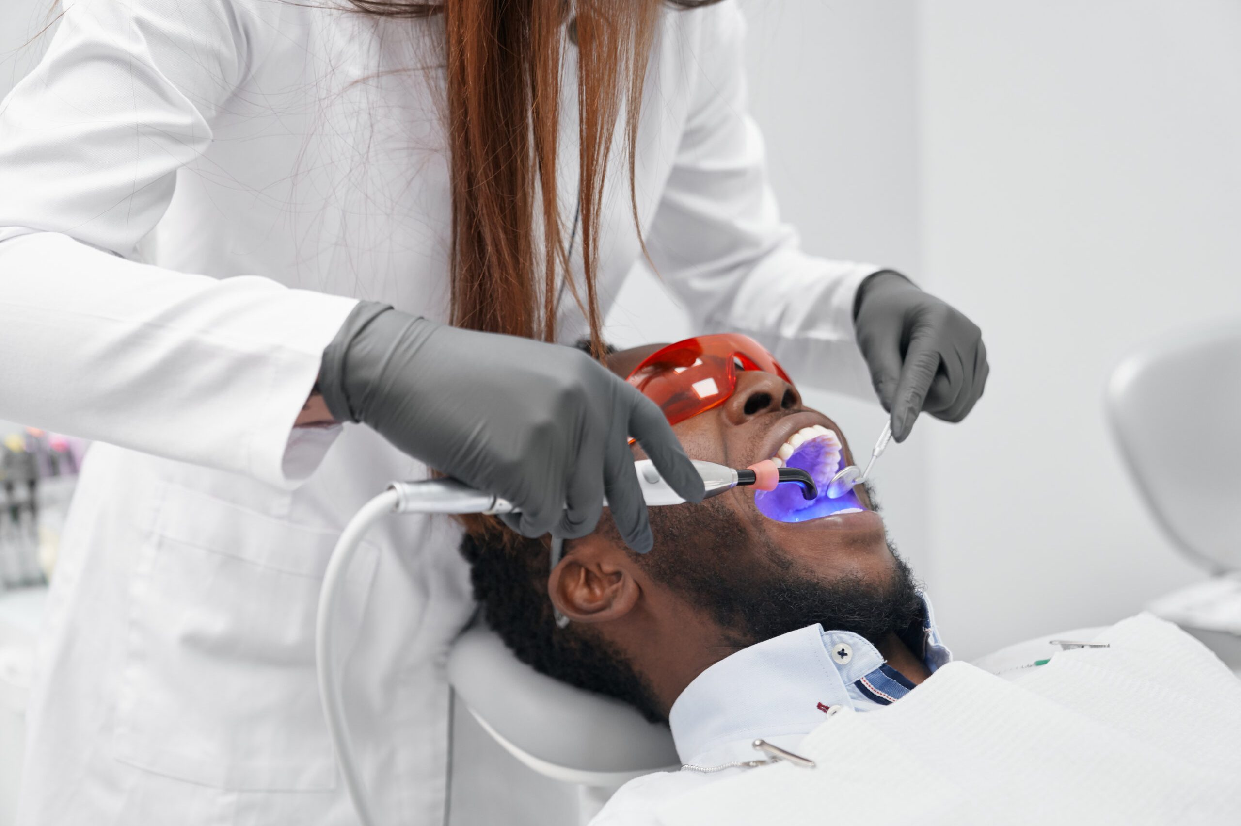View from side of female doctor in process of doing filling to african man. Male patient lying with open mouth while doctor keeping ultraviolet device. Concept of stomatology and care.